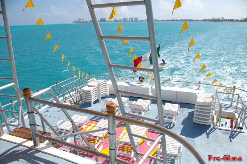 Cubierta superior del catamarán Dancer con tumbonas blancas y banderines amarillos sobre el agua turquesa y la silueta de Cancún