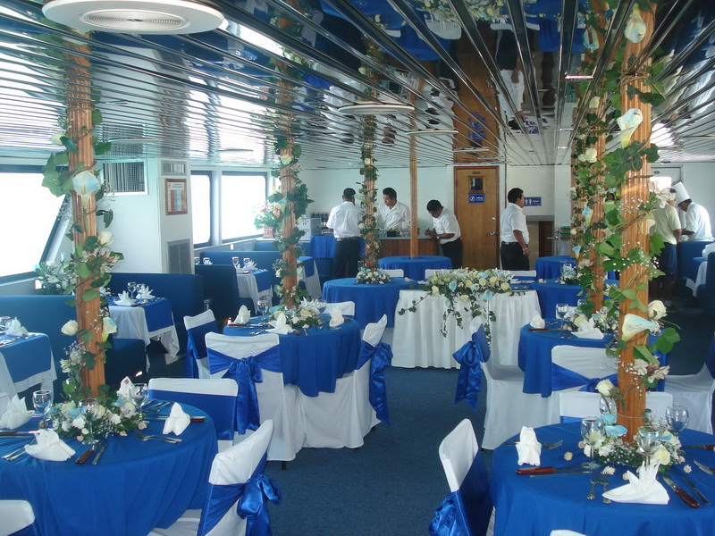 Salón inferior del barco Discovery montado como recepción de boda con manteles azul rey, sillas vestidas de blanco y columnas florales