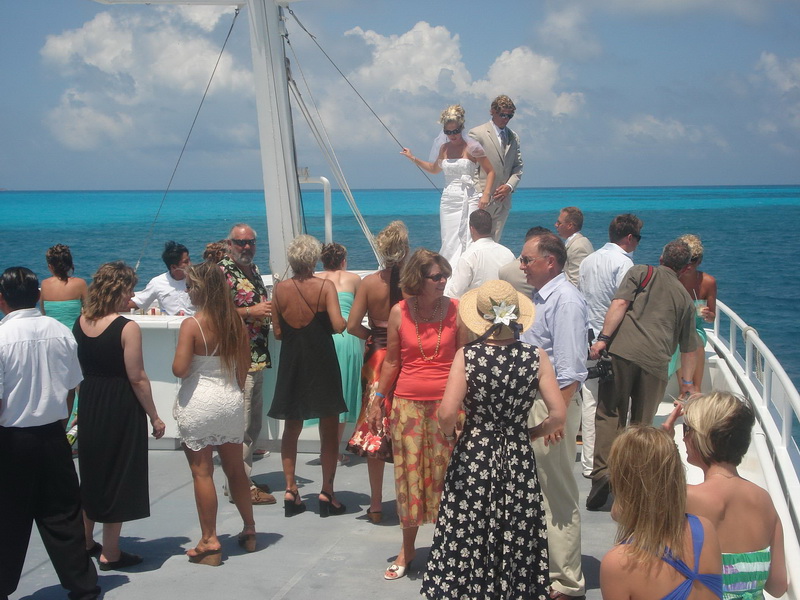 Novios de pie en la proa del barco Discovery rodeados de invitados con el agua turquesa brillante del Caribe alrededor
