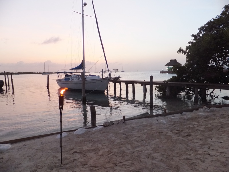 Velero anclado al atardecer junto al muelle y la playa de arena de Sunset Grill Isla Mujeres, con palapa y antorcha junto a la laguna en calma