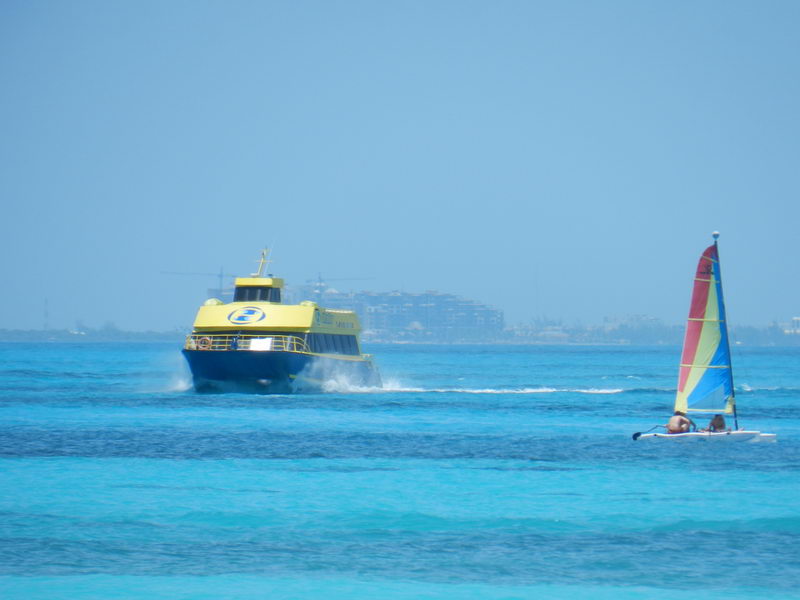 Yellow Ultramar ferry crossing to Isla Mujeres past a colorful Hobie Cat sailboat over bright turquoise water